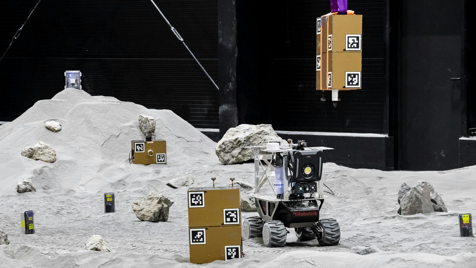 An small uncrewed vehicle traverses a lunar-like surface that has gold-colored boxes with symbols on them.