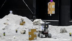 An small uncrewed vehicle traverses a lunar-like surface that has gold-colored boxes with symbols on them. An small uncrewed vehicle traverses a lunar-like surface that has gold-colored boxes with symbols on them.