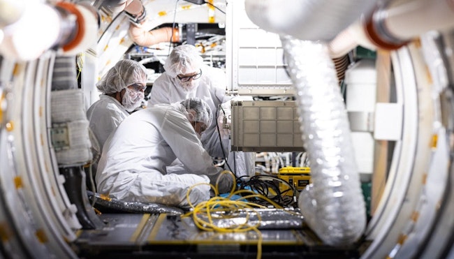 A trio of people in cleanroom suits work with electronics.