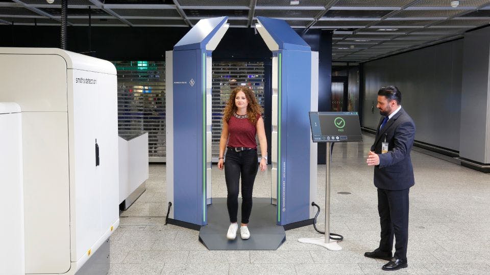 A woman walks through an airport screening device as a man directs her to walk through as a green check mark appears on his screen.