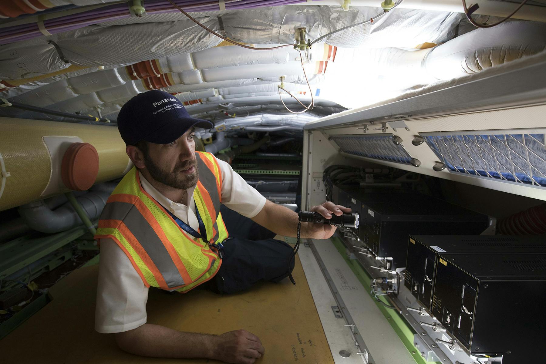 A tech service worker uses a flashlight to inspect equipment in an airplane.