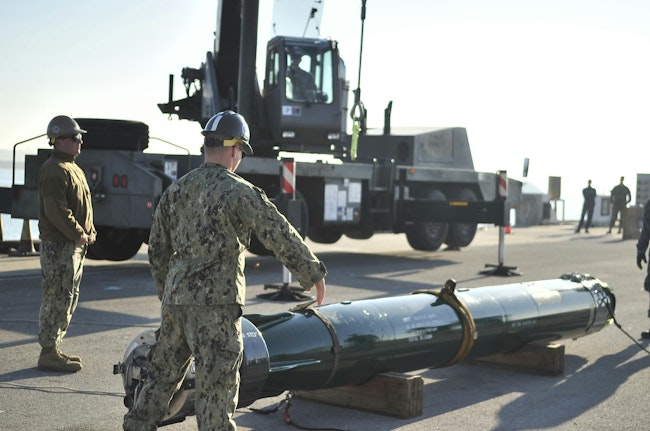 A U.S. Navy equipment operator signals the crane to lower the hook to attach an MK 48 inert training torpedo during an expeditionary ordnance loading exercise.