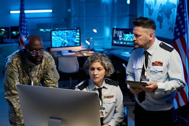 Team of serious workers of military command center looking at online data while sitting and standing in front of computer monitor