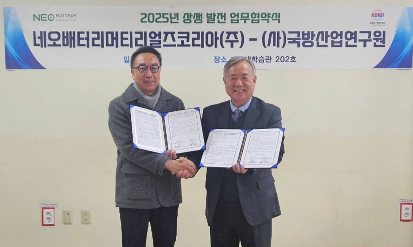 Two men shake hands in front of a Korean-language backdrop.