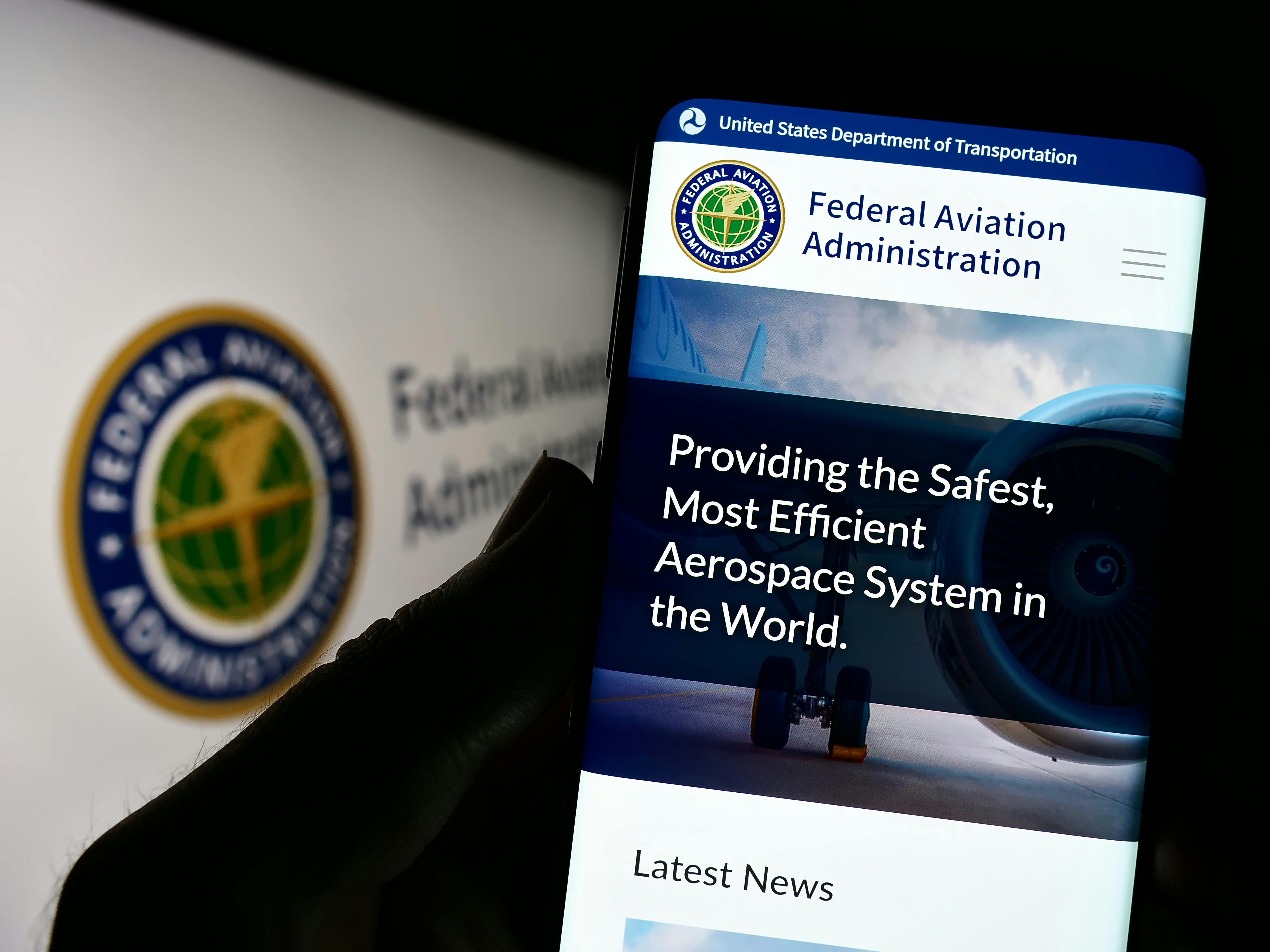 Person holding smartphone with website of US Federal Aviation Administration (FAA) on screen in front of seal. Focus on center of phone display.