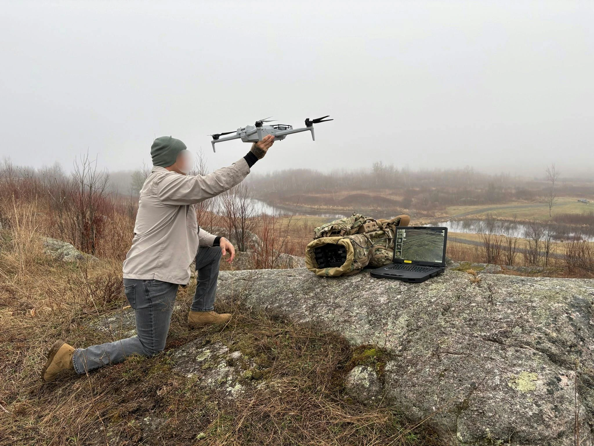 A person with their face obscured assists a drone in taking off from a ridge. A laptop sits on a rock beside a bag.