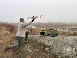 A person with their face obscured assists a drone in taking off from a ridge. A laptop sits on a rock beside a bag. A person with their face obscured assists a drone in taking off from a ridge. A laptop sits on a rock beside a bag.