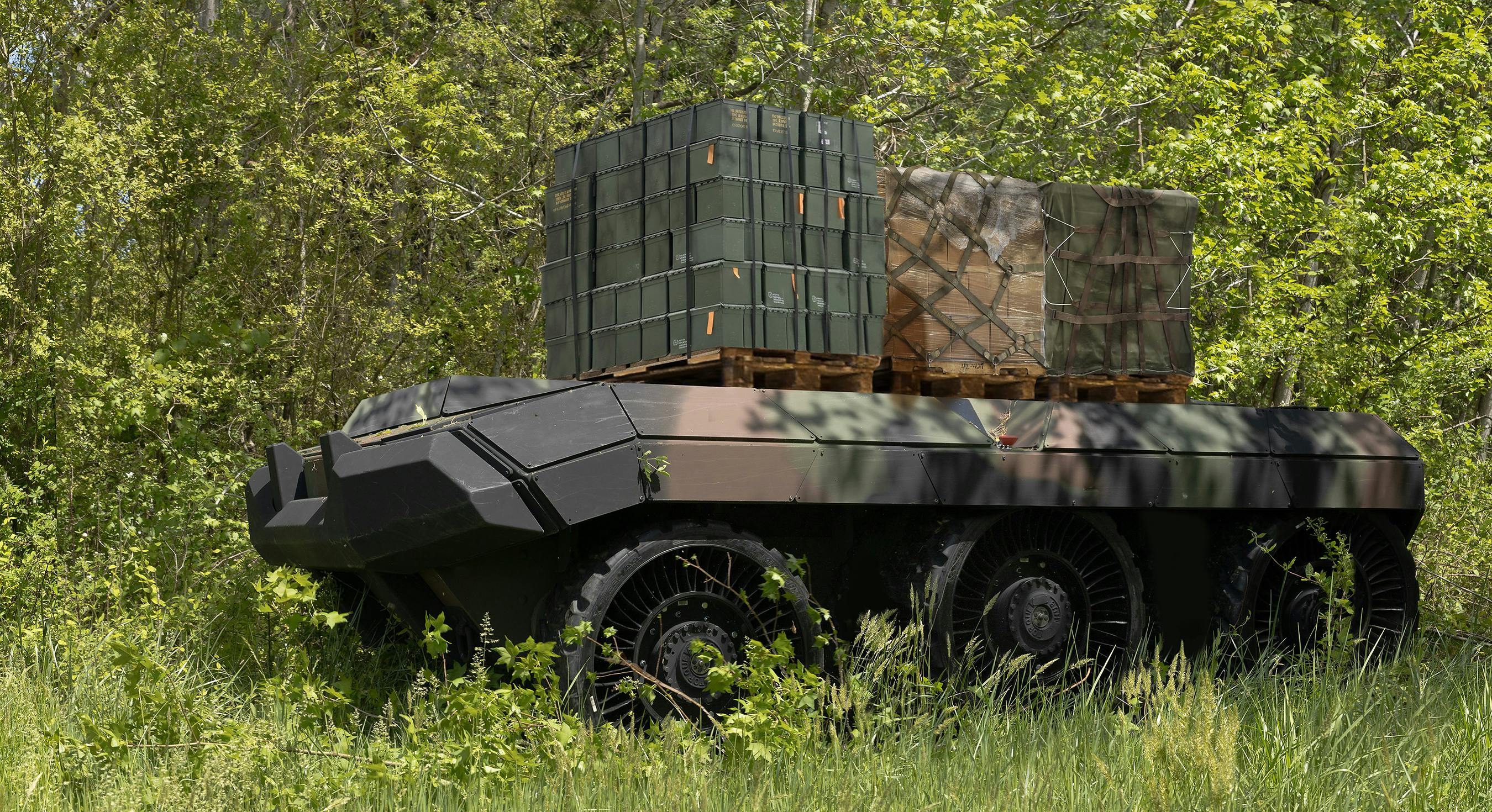 A six-wheeled, flat-topped uncrewed vehicle carries ammo containers and other materiel in a grassy field