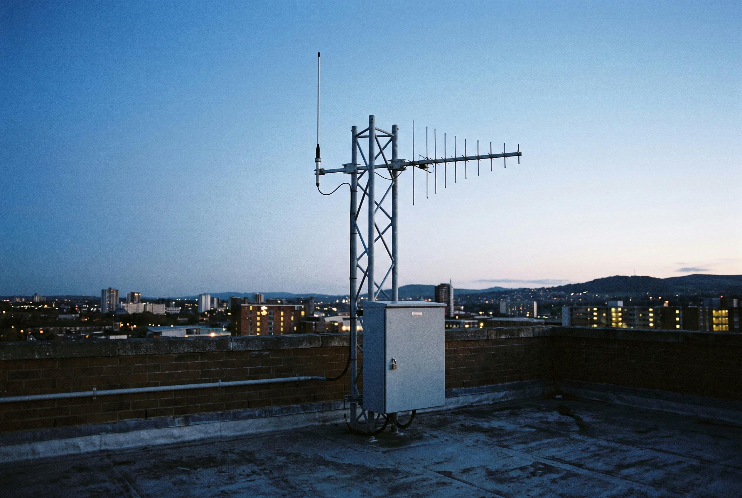 A rooftop monitoring site: an omnidirectional antenna and a log-periodic antenna on a lattice mast, with the equipment cabinet at its base. No real site is depicted.