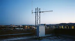 A rooftop monitoring site: an omnidirectional antenna and a log-periodic antenna on a lattice mast, with the equipment cabinet at its base. No real site is depicted.