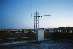 A rooftop monitoring site: an omnidirectional antenna and a log-periodic antenna on a lattice mast, with the equipment cabinet at its base. No real site is depicted.