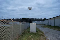A site perimeter: a fence line, a slim mast carrying two panel antennas and a dome, an equipment cabinet, and a small quadcopter in the distance. No real site is depicted.